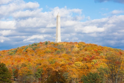A tall monument stands atop a hill surrounded by vibrant autumn foliage under a cloudy sky.