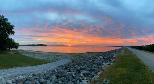 A serene lakeside view at sunset, with colorful clouds reflecting on the water and a rocky shoreline.