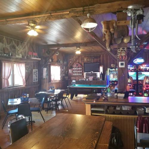 Interior of a rustic bar with wooden decor, tables, and a pool table, featuring dim lighting and a cozy atmosphere.