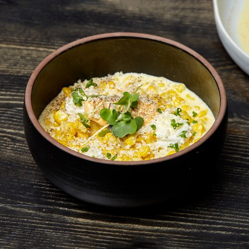A bowl of creamy corn dish topped with microgreens and spices, set on a dark wooden surface.