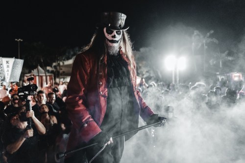 A performer in a red coat and top hat stands over a crowd, surrounded by smoke and dramatic lighting at night.