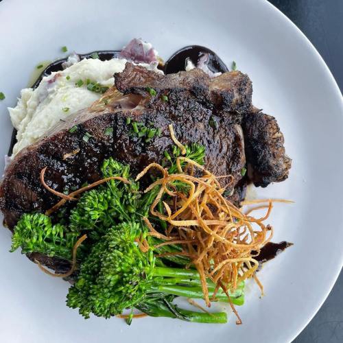 A plate featuring a grilled steak, creamy mashed potatoes, broccolini, and crispy fried onions drizzled with sauce.
