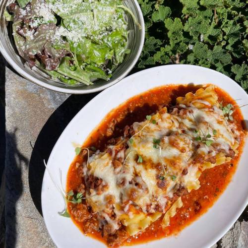 A plate of lasagna topped with cheese and herbs, served with a side salad in a bowl, set on a stone surface.