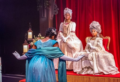 A theatrical scene featuring three women in elaborate historical costumes, with one performing in front of two seated figures.