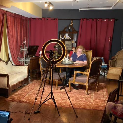 A woman sits at a table with a camera and ring light, surrounded by red curtains and vintage furniture in a cozy room.