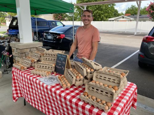 A smiling vendor stands behind a table of fresh eggs in cartons at a market, with a checkered tablecloth and green canopy.