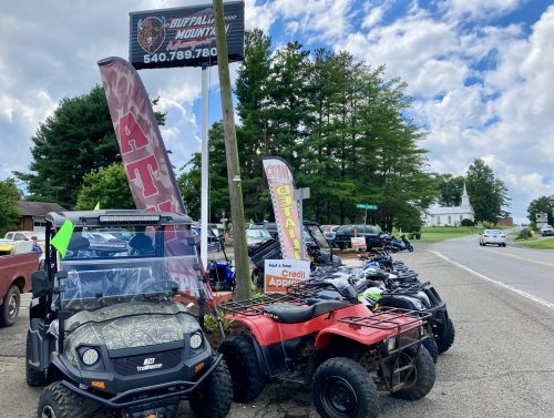 A lineup of ATVs and a utility vehicle for sale outside a dealership, with trees and a road in the background.