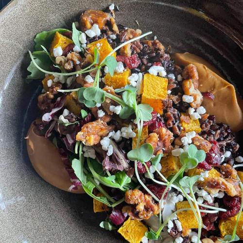 A colorful salad with mixed greens, roasted squash, walnuts, cranberries, and goat cheese on a dark plate.
