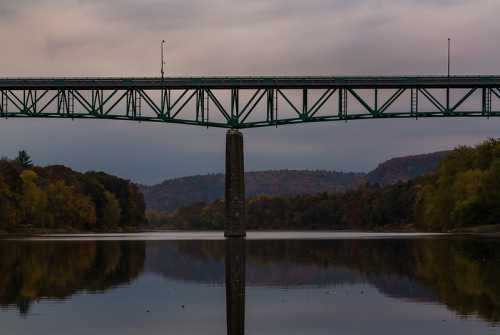 A green bridge spans a calm river, reflecting autumn trees and a cloudy sky in the background.