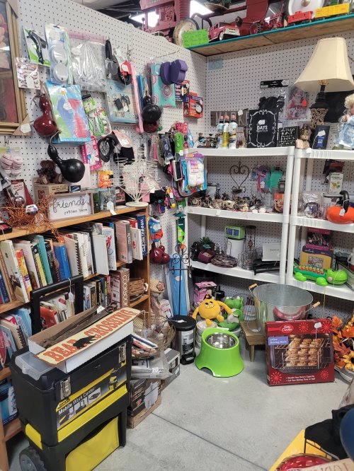 A cluttered thrift store display featuring various household items, books, and decorative objects on shelves.