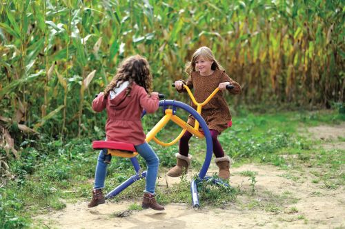Two young girls play on a colorful outdoor seesaw in a cornfield, smiling and enjoying their time together.