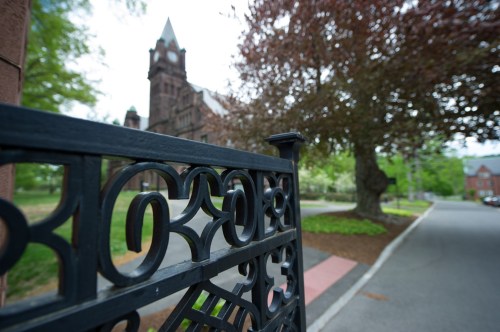 A decorative black gate in the foreground with a historic building and trees in the background on a quiet street.