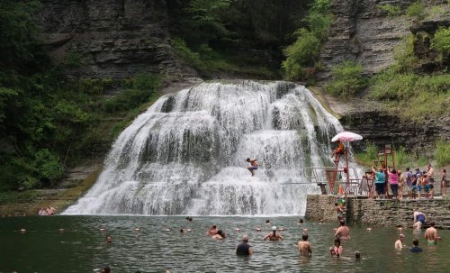 A waterfall cascades into a swimming area where people are enjoying the water and sunbathing.