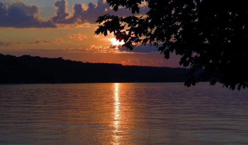 A serene sunset over a calm lake, with vibrant colors reflecting on the water and silhouetted trees in the foreground.