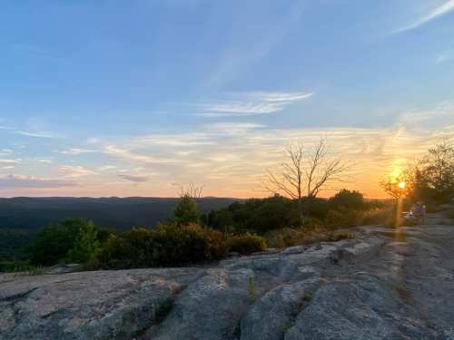 A scenic view of a sunset over a mountainous landscape, with rocky foreground and trees silhouetted against the sky.
