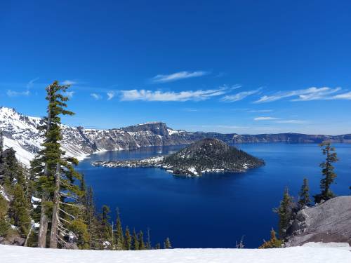 A panoramic view of Crater Lake, featuring a deep blue lake surrounded by snow-capped mountains and evergreen trees.