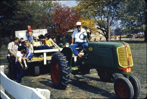 A man drives a green tractor with children on a hayride in a rural setting, surrounded by trees in autumn colors.