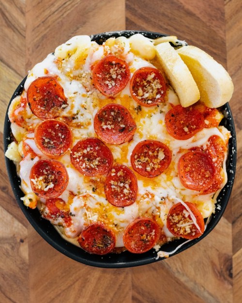 A bowl of baked macaroni and cheese topped with pepperoni slices and breadcrumbs, served with a slice of bread.