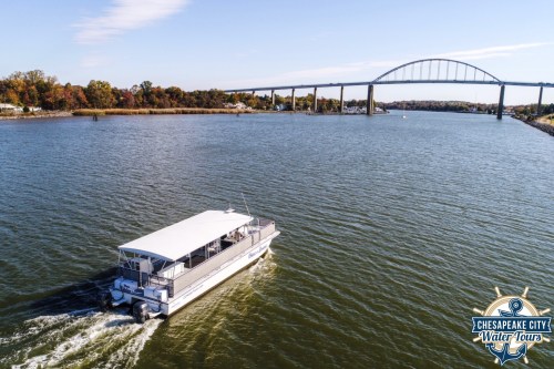 A boat navigates a calm river under a bridge, surrounded by autumn-colored trees. Chesapeake City Water Tours logo in corner.