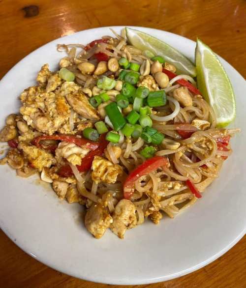 A plate of stir-fried noodles with chicken, peanuts, red peppers, green onions, and lime wedges.