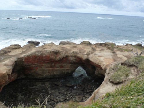 A rocky coastal arch overlooking the ocean, with waves crashing against the shore under a cloudy sky.
