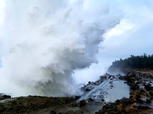 A powerful wave crashes against rocky shore, sending up a spray of white foam against a cloudy sky.