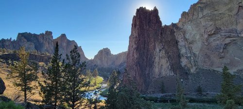 A scenic view of towering rock formations and a river, with sunlight shining over the peaks in a clear blue sky.