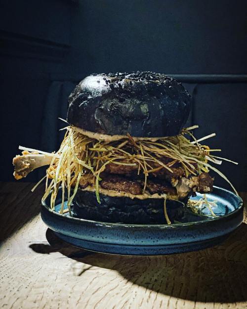A dark burger with a black bun, layered with crispy fried onions and a chicken drumstick, served on a blue plate.