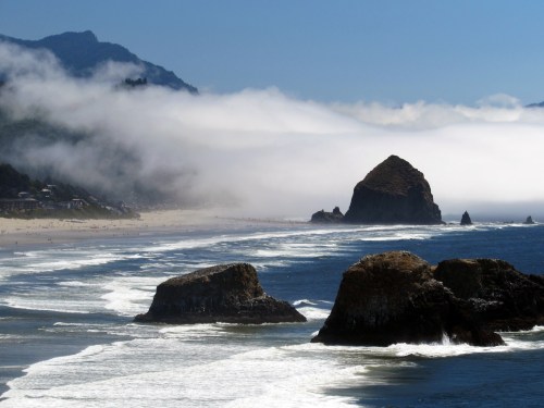 A scenic coastal view with rocky formations, gentle waves, and fog rolling over the beach and mountains in the background.
