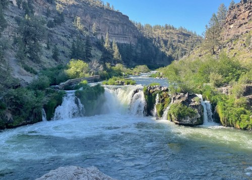 A serene river flows over a waterfall, surrounded by lush greenery and rocky cliffs under a clear blue sky.