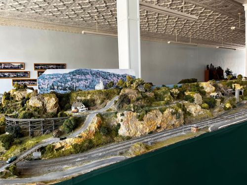 A detailed model landscape featuring hills, trees, and miniature buildings, with train tracks winding through the scene.