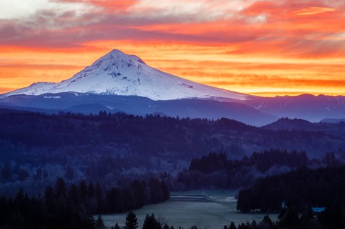Snow-capped mountain at sunset, with vibrant orange and purple skies above a forested landscape.