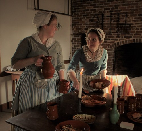 Two women in historical attire serve food at a rustic table, illuminated by candlelight in a cozy, brick-walled setting.