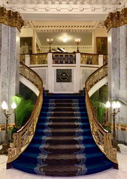 Elegant staircase with a blue carpet, ornate railings, and marble walls, leading to a grand landing with artwork.