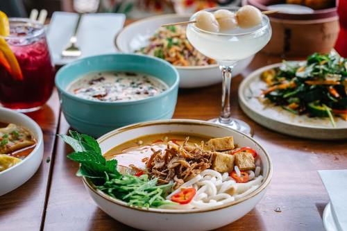 A bowl of noodles topped with herbs, tofu, and chili, alongside drinks and other dishes on a wooden table.