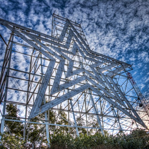 A large, metallic star structure against a blue sky with clouds, surrounded by greenery.