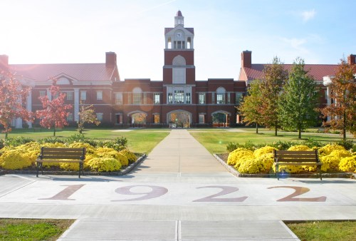 A sunny campus scene featuring a brick building, landscaped gardens, and benches along a pathway marked "1922."
