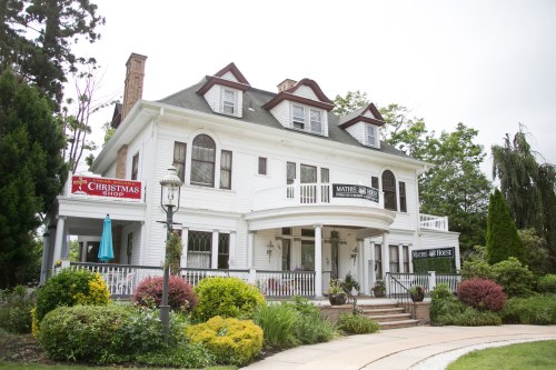 A large white Victorian-style house with a wraparound porch, surrounded by colorful gardens and signage for a Christmas shop.