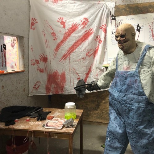 A dimly lit room with a blood-stained wall, a creepy figure in overalls, and unsettling props on a table.