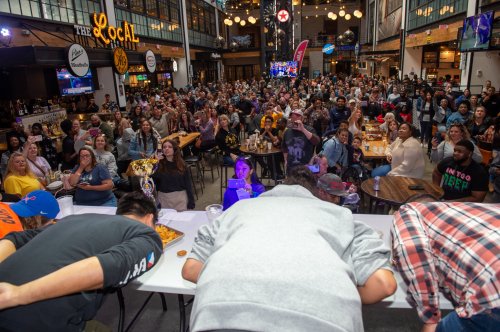 A large crowd watches a competitive eating event in a lively indoor venue, with participants leaning over a table.
