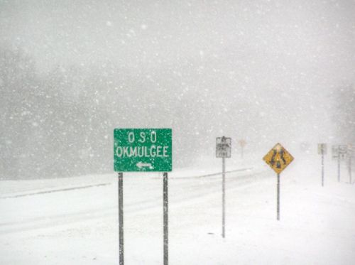 Snowy scene with a green road sign for Oso and Okmulgee, surrounded by heavy snowfall and blurred road signs.