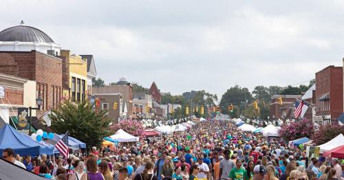 A bustling street festival with large crowds, colorful tents, and decorations lining the road under a cloudy sky.