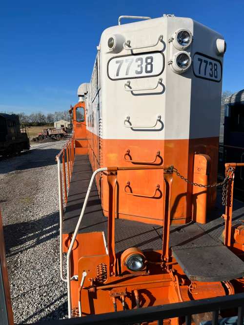 A close-up of an orange and white vintage train engine, numbered 7738, against a clear blue sky.