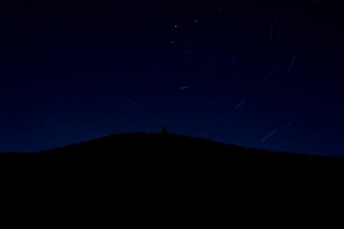 A dark silhouette of a hill against a starry night sky with star trails creating circular patterns.