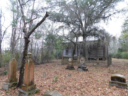 An old, abandoned house surrounded by trees and a graveyard with weathered tombstones, covered in fallen leaves.