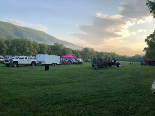 A grassy field with parked vehicles, tents, and mountains in the background under a cloudy sky.