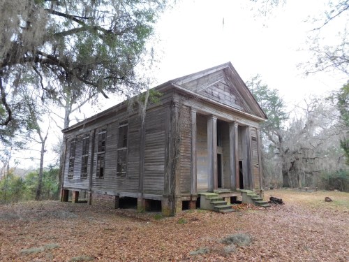 An old, abandoned wooden building with a classic façade, surrounded by trees and fallen leaves.