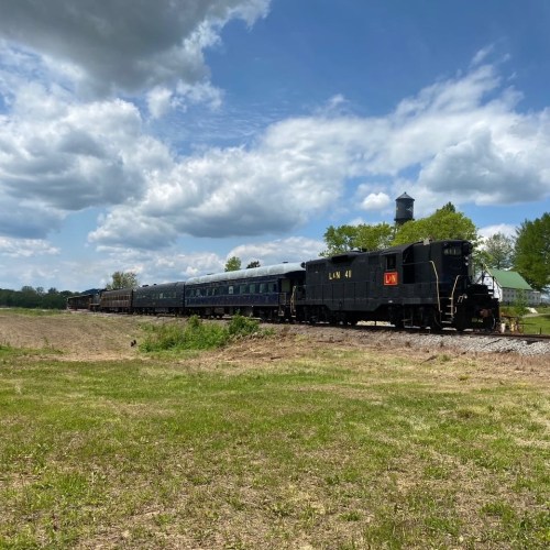 A vintage black train engine on tracks, surrounded by green fields and a cloudy blue sky.