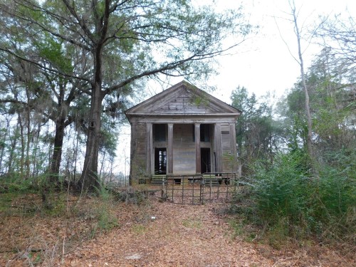 Abandoned wooden house surrounded by trees and overgrown vegetation, with a path leading to a gated entrance.
