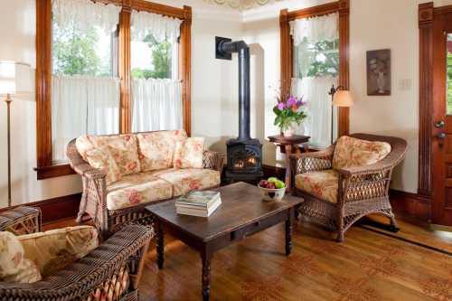 Cozy living room with floral furniture, a wooden table, and a black stove, surrounded by bright windows.